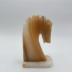May include: A carved stone horse head sculpture in shades of brown and cream. The sculpture has a curved neck and a detailed mane. The base is a rectangular shape. The horse head is a decorative item.