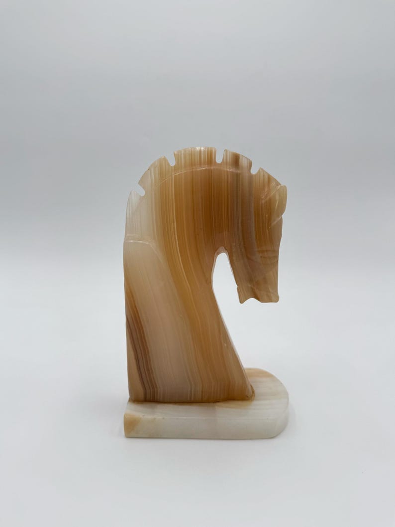 May include: A carved stone horse head sculpture in shades of brown and cream. The sculpture has a curved neck and a detailed mane. The base is a rectangular shape. The horse head is a decorative item.