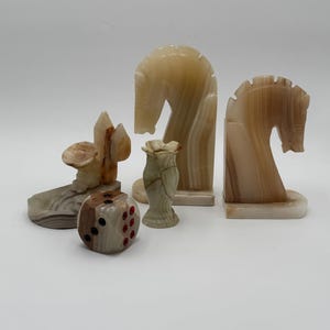 Pode incluir: Uma coleção de itens decorativos esculpidos em ônix. Os itens incluem dois suportes de livros com cabeça de cavalo, um vaso pequeno, um dado e uma escultura de uma pessoa com um chapéu. Os itens são em tons de bege, marrom e creme.