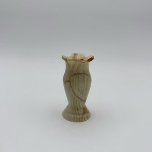 May include: A small, decorative vase made of a light green and brown stone. The vase has a flared top and a rounded base. The stone has natural veining and a polished finish. The vase is approximately 6 inches tall.