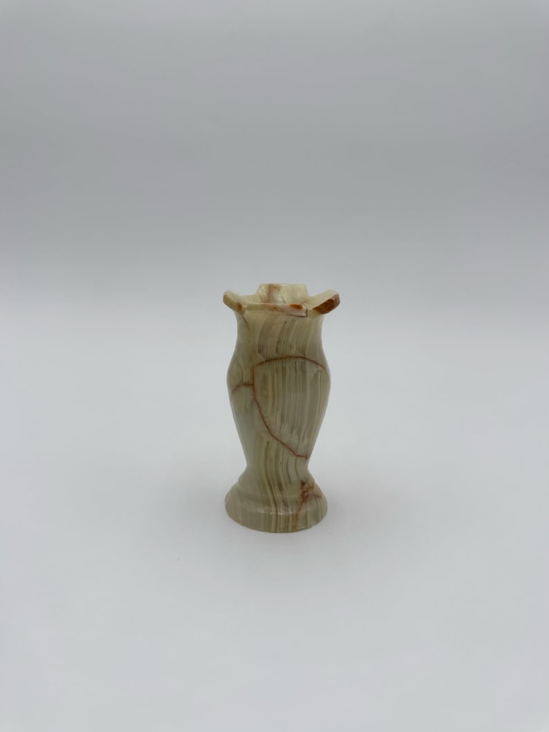 May include: A small, decorative vase made of a light green and brown stone. The vase has a flared top and a rounded base. The stone has natural veining and a polished finish. The vase is approximately 6 inches tall.