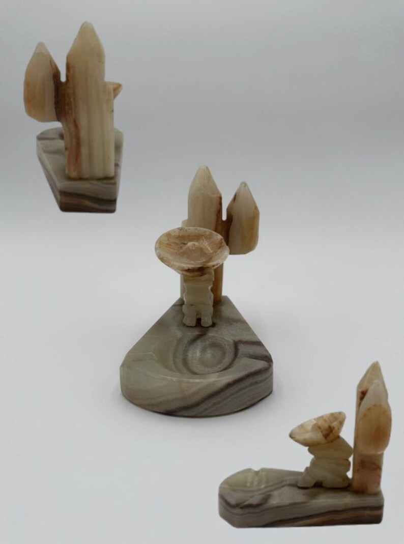 May include: Three carved onyx figurines. One depicts a cactus, another a man in a sombrero, and the third a man with a cactus. The figurines are in shades of beige, brown, and gray, with a polished finish. The bases are rectangular or triangular.