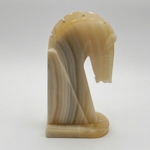 May include: A decorative horse sculpture in shades of beige and tan. The art piece features a stylized horse head with a flowing mane and a curved neck. The sculpture is made of a stone-like material and has a smooth, polished finish.