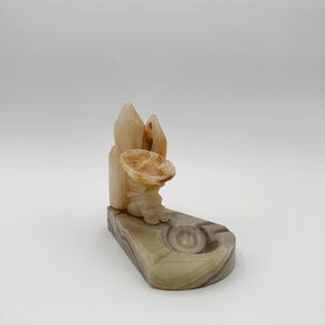 May include: A carved stone ashtray with a sombrero-shaped design. The ashtray is made of beige and brown stone, with a textured surface. The design includes a sombrero and a decorative element.