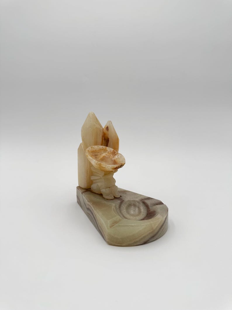 May include: A carved stone ashtray with a sombrero-shaped design. The ashtray is made of beige and brown stone, with a textured surface. The design includes a sombrero and a decorative element.