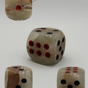 May include: A collection of stone dice with a marbled appearance. The dice have a light green and brown color scheme, with black and red dots indicating the numbers. The dice are shown from multiple angles.
