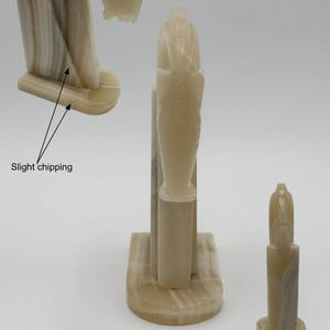 May include: A collection of beige and cream-colored onyx sculptures. The sculptures feature a stylized, abstract design with vertical and curved elements. One sculpture shows slight chipping. The pieces are displayed against a white background.