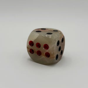 May include: A single, beige-colored dice with red and black dots. The dice is made of a translucent material, with visible brown and tan veining. The dots are evenly spaced on each face, indicating the numbers one through six.
