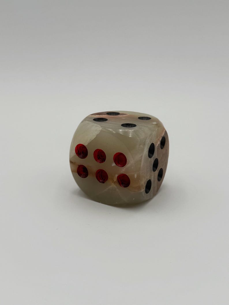 May include: A single, beige-colored dice with red and black dots. The dice is made of a translucent material, with visible brown and tan veining. The dots are evenly spaced on each face, indicating the numbers one through six.