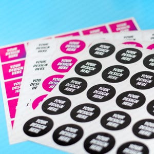 Può includere: Fogli di adesivi con la scritta "YOUR DESIGN HERE" in bianco e nero su sfondo bianco. Alcuni adesivi sono rotondi con un cerchio nero, altri rettangolari con sfondo rosa. Gli adesivi sono su una superficie azzurra.