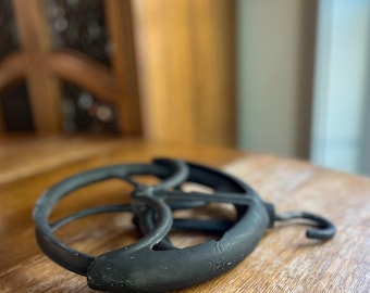 May include: A black cast iron pulley with a curved body and a hook. The pulley is resting on a wooden surface. The pulley's wheel is visible, and the overall design suggests an antique or vintage item.