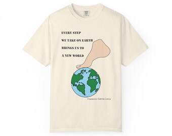 May include: Cream-colored t-shirt featuring a graphic of a foot pressing down on a globe. The text reads "EVERY STEP WE TAKE ON EARTH BRINGS US TO A NEW WORLD" with "Frederico Garcia Lorca" below. The design is in black, green, blue, and skin tone.