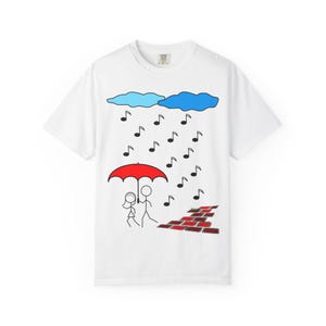 May include: White t-shirt with a whimsical design featuring a red umbrella sheltering two stick figures from musical notes falling from blue clouds. A brick wall is on the right side of the design.