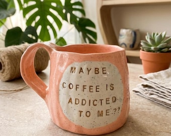 May include: A peach-colored ceramic mug with a speckled finish and a wavy, organic shape. The front features a circular cream-colored patch with the text MAYBE COFFEE IS ADDICTED TO ME?? in dark brown, stamped-style lettering.