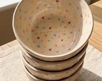 May include: A stack of three off-white ceramic bowls with a glossy finish. The interior of the top bowl features small, hand-painted multicolored hearts and the word HAPPINESS stamped in the center. The text MONETA CERAMIC appears in the corner.