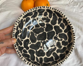 May include: A ceramic bowl with a black and white cow print design. The bowl has a cream-colored base with irregular black shapes. The rim is decorated with small black dots. Three oranges are in the background.
