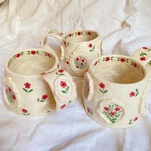 May include: Three cream-colored ceramic mugs featuring hand-painted red flowers with green stems and leaves. Each mug is adorned with raised, ornate, oval-shaped frames containing floral motifs, and the rims are decorated with small red flower patterns.