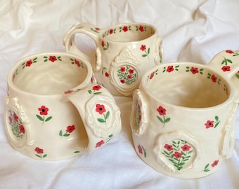 May include: Three cream-colored ceramic mugs featuring hand-painted red flowers with green stems and leaves. Each mug is adorned with raised, ornate, oval-shaped frames containing floral motifs, and the rims are decorated with small red flower patterns.