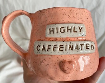 May include: A speckled peach-colored ceramic mug with the words "HIGHLY CAFFEINATED" in white lettering. The mug has a heart-shaped detail and a large handle. The mug is designed for hot beverages.