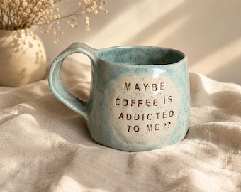 May include: A light blue ceramic mug with a speckled finish and a large, curved handle. The front features a circular white area with the embossed text: MAYBE COFFEE IS ADDICTED TO ME?? The mug has an irregular, organic shape and a glossy surface.