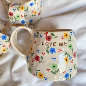 Peut inclure: Une tasse en céramique de couleur crème avec un motif floral peint à la main composé de petites fleurs rouges, bleues, jaunes et vertes sur des tiges vertes. La tasse présente une surface texturée irrégulière et le texte doré I LOVE ME.