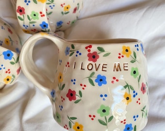 May include: A cream-colored ceramic mug with a hand-painted floral pattern featuring small red, blue, yellow, and green flowers with green stems. The mug has a textured, uneven surface and the gold-colored text I LOVE ME printed on the front.