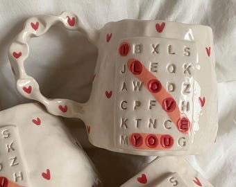 May include: White ceramic mugs with red heart accents and handles. One mug features a crossword puzzle design with the word "LOVE" highlighted in red. The mugs are handmade and perfect for coffee or tea.