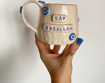 May include: A cream-colored ceramic mug with an uneven, hand-molded texture. The mug features blue evil eye beads attached to the surface and handle. The text "SAY" and "MASALLAH" is embossed in blue lettering on the front of the mug.