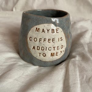 Peut inclure: Une tasse en céramique bleu-gris faite à la main avec le texte "MAYBE COFFEE IS ADDICTED TO ME??" dans un cercle blanc. La tasse a une forme irrégulière et une texture mouchetée, parfaite pour le café ou le thé.