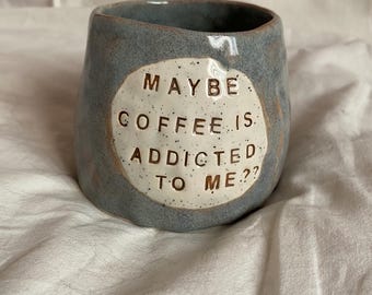 May include: A handmade, blue-grey ceramic mug with the text "MAYBE COFFEE IS ADDICTED TO ME??" in a white circle. The mug has a unique, irregular shape and a speckled texture, perfect for coffee or tea.