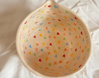May include: A cream-colored ceramic bowl with a heart-shaped design in various colors. The word "HAPPINESS" is written on the side of the bowl. The bowl is set against a white background.