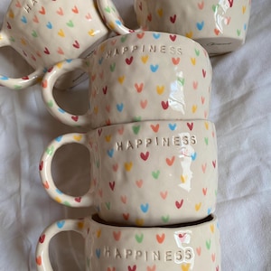 Könnte beinhalten: Eine Sammlung von fünf cremefarbenen Keramikbechern mit einer strukturierten, genoppten Oberfläche. Jeder Becher trägt das Wort HAPPINESS in Prägung nahe dem Rand und ist mit einem wiederkehrenden Muster aus kleinen, bunten Herzen in Rot, Blau, Gelb und Grün verziert.