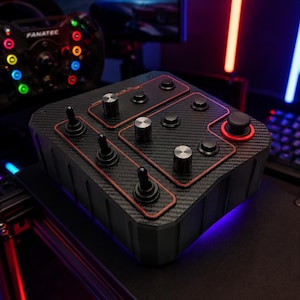 Op de afbeelding: Een zwarte knoppenbox met een koolstofvezeltextuur, meerdere metalen draaiknoppen, tuimelschakelaars en drukknoppen. Het apparaat heeft rode accentlijnen rond de bedieningspanelen en een enkele joystickknop met een rode rand.