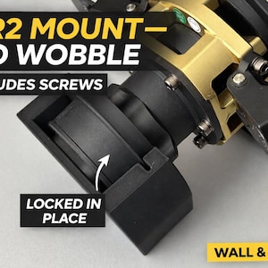 Op de afbeelding: Close-up van een zwarte en gouden QR2-bevestiging met de tekst "QR2 MOUNT - NO WOBBLE" en "INCLUDES SCREWS". De bevestiging is ontworpen om op zijn plaats te vergrendelen en is voor een muur en sim rig.