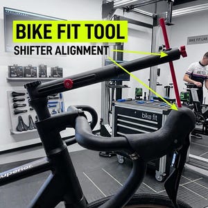 May include: A black bike fit tool for shifter alignment is positioned on a bicycle handlebar. The tool features a horizontal black bar with a red adjustment knob and a vertical red rod that extends downward to align with the bike shifter.