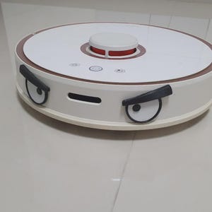 May include: A white and brown robotic vacuum cleaner with a circular design. The top features a white center with a red accent and a brown trim. The front has cartoon-like eyes. This automated cleaner is designed for floor cleaning.