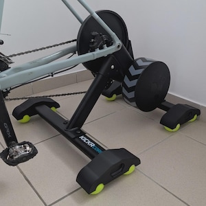 Op de afbeelding: Een zwarte en grijze fietstrainer met een lichtblauw fietsframe. De trainer heeft zwarte steunen met gele wielen en de tekst "KICKR CORE". De trainer heeft een zwart vliegwiel met grijs chevron-ontwerp.