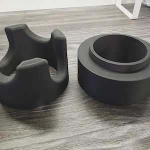 Op de afbeelding: Twee donkergrijze 3D-geprinte objecten. Eén is een wiegachtige vorm met gebogen steunen. De andere is een cilindrische vorm met een verhoogde binnenring. Beide objecten staan op een lichtgrijze ondergrond.
