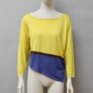 May include: A long-sleeved top in yellow, blue, and brown. The top has a boat neck and a color-blocked design. The top is displayed on a mannequin. The yellow portion covers the top half, with a brown stripe separating it from the blue bottom.