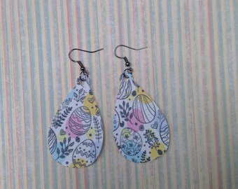 May include: A pair of dangle earrings with a teardrop shape. The earrings are white with a pastel yellow, blue, and pink pattern of Easter eggs. The earrings have silver hooks.