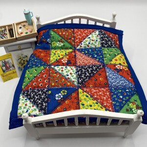 May include: Miniature white bed with a colorful patchwork quilt. The quilt features red, blue, green, and yellow triangles with floral and dotted patterns. A small nightstand with art supplies and a watercolor book is next to the bed.