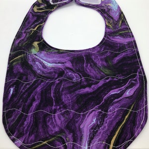 May include: Purple and gold marbled baby bib with white stitching. The bib has a scalloped edge and a hook and loop closure.