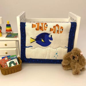 May include: A miniature white crib with a blue and white blanket featuring cartoon fish. A small brown teddy bear with a pink bow sits nearby. A miniature nightstand holds a lamp, a stack of books, and a basket of books.