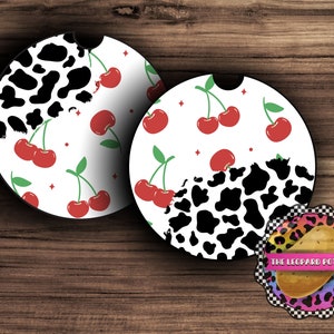Op de afbeelding: Twee ronde onderzetters met een zwart-witte koe-print ontwerp en rode kersen op een witte achtergrond. De onderzetters liggen op een houten oppervlak. De tekst "THE LEOPARD POTATO" staat op een kleinere onderzetter met een luipaardprint.