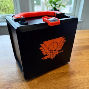 May include: A black storage box with an orange handle and latch. The box has a skull and crossbones logo with wings on the front.