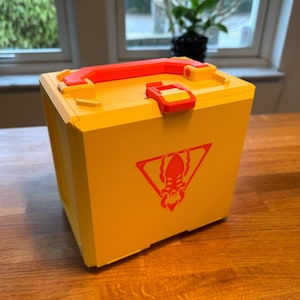 May include: A yellow plastic storage box with a red handle and latch. The box has a red logo of a creature with a triangular shape around it.