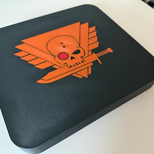 Peut inclure: Un dessous de verre carré noir avec un motif orange de crâne et d'épée. Le crâne a un œil rouge et est entouré d'ailes.