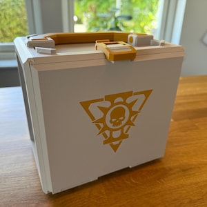 May include: A white box with a yellow handle and clasp. The box features a yellow emblem with a skull and sunburst design. The box is sitting on a wooden surface.