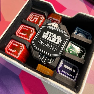 Pode incluir: Uma caixa branca com inserções de espuma preta contém vários dados coloridos com letras brancas. Os dados são etiquetados com números e símbolos. Um token preto com as palavras "Star Wars Unlimited" está no centro da caixa.
