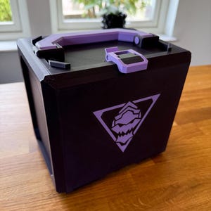May include: A black box with a purple handle and accents. The box features a purple geometric design on the front. The box is sitting on a wooden surface.